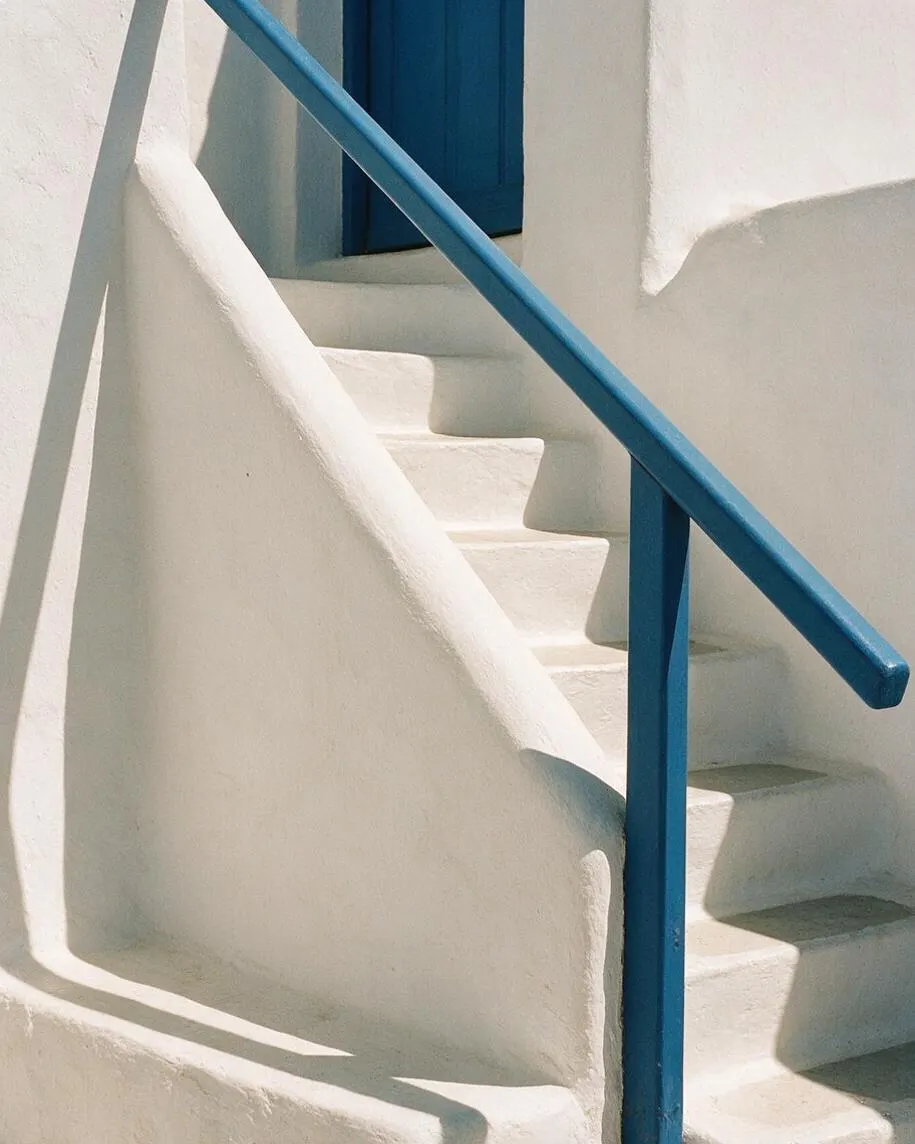 A Mediterranean-style exterior staircase with smooth white stucco walls and built-in steps, accented by a vivid cobalt blue handrail and matching door shutters. Strong sunlight creates crisp shadows across the curving surfaces, emphasizing the simple, sculptural architecture.