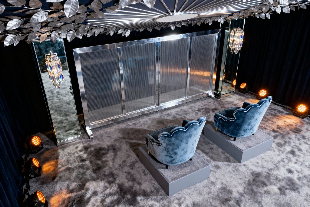 Media room with two dusty-blue velvet chairs on gray plinths facing a wall clad in fluted glass within a polished chrome frame. Mirrored columns flank the wall, each holding a crystal prism sconce. Deep navy velvet curtains surround the space as amber spotlights wash the base. The floor is high-pile gray carpet with fur-like texture, and a silver sunburst ceiling edged with metal leaves encircles a central round light.