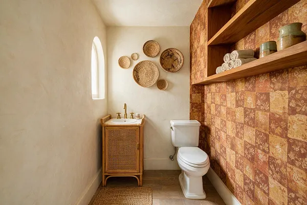 A small powder room with textured cream walls, a narrow arched window, and a woven rattan vanity with a brass faucet and white sink. Woven wall baskets decorate the back wall, while patterned terracotta-toned wallpaper, open wooden shelves, and a white toilet add warmth and character.