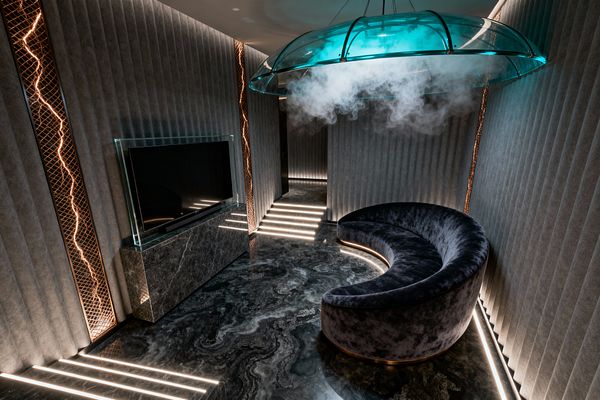 A futuristic media room with a crescent-shaped charcoal velvet sofa on a glossy black-and-gray stone floor with swirling, galaxy-like veining. Fluted gray wall panels are interrupted by copper lattice vertical insets lit like lightning. A large flat-screen sits on a dark marble console with a clear glass frame. Linear LED strips outline steps and baseboards. Overhead, a translucent teal dome pendant releases mist, bathing the space in a blue-green glow.
