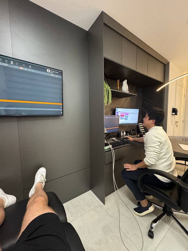 A person is seated at a desk in a modern office environment, working on a computer while another person relaxes in a chair.