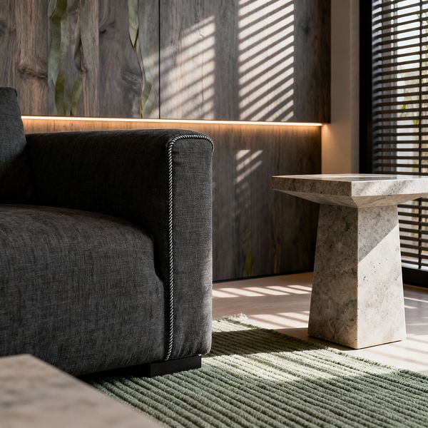 A close-up of a modern living room shows a charcoal-gray upholstered sofa with white contrast piping on a ribbed sage green rug. A light gray honed stone pedestal side table sits nearby, while vertically grained gray wood wall panels and a slim recessed light create a warm glow as sunlight through dark slatted blinds casts striped shadows across the floor and surfaces, highlighting clean lines and tactile neutral materials.