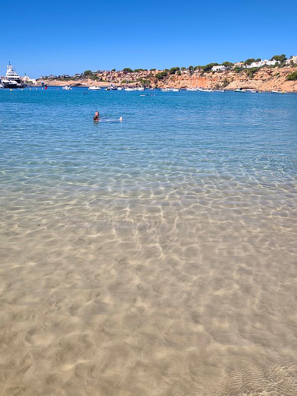 A person swimming in clear blue water near a sandy beach with boats and a rocky coastline in the background under a clear sky.