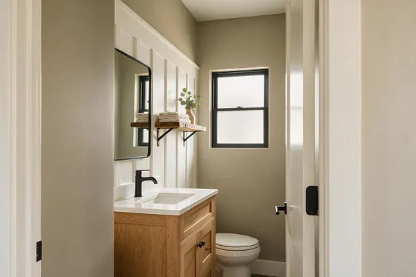 A narrow farmhouse-style powder room with a light oak vanity, white countertop, matte black faucet, and framed mirror. Taupe walls, a black-trimmed window, and a floating wood shelf with folded towels and greenery create a warm, tidy feel.