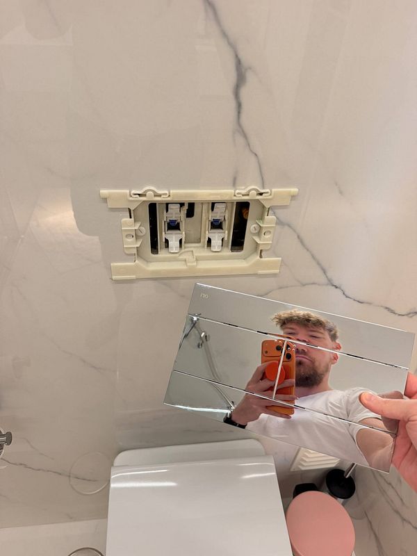 Toilet flush system installed upside down on a wall with the flush button panel removed and held in hand, reflected showing the person taking the photo.