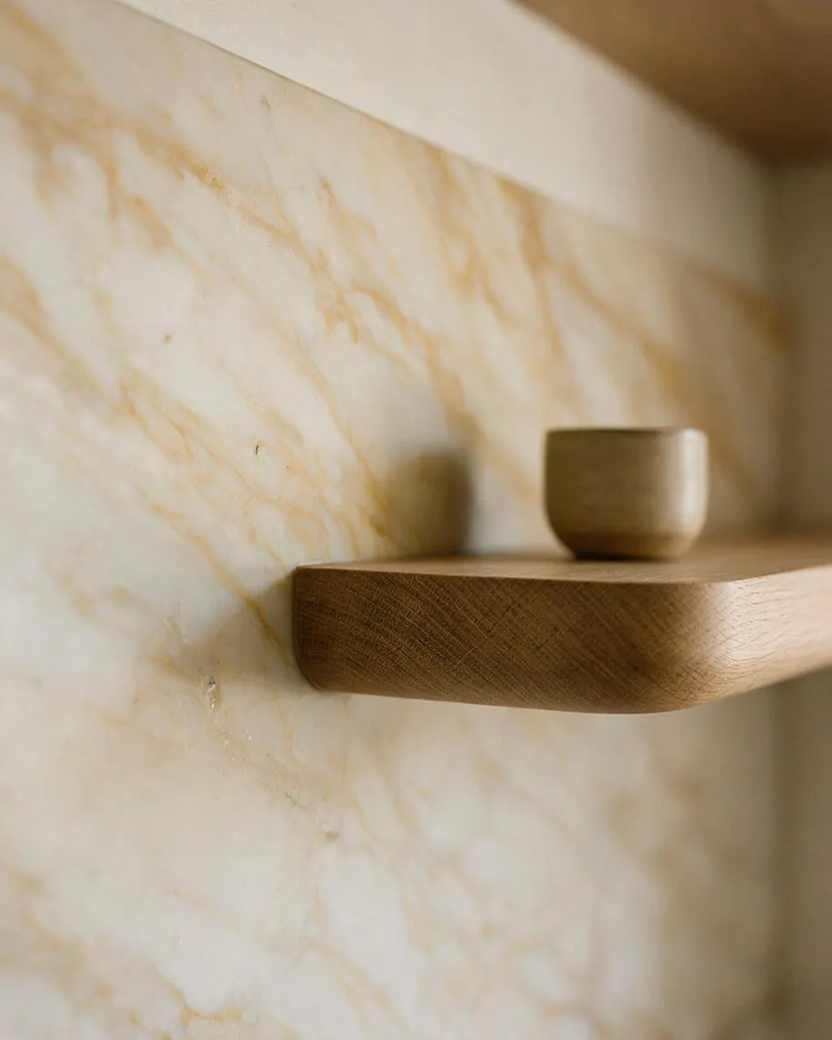 A close-up interior detail shows a rounded floating wooden shelf mounted against a cream marble wall with soft gold veining. A small matching cup sits on the shelf, creating a calm, minimal composition with warm natural materials.