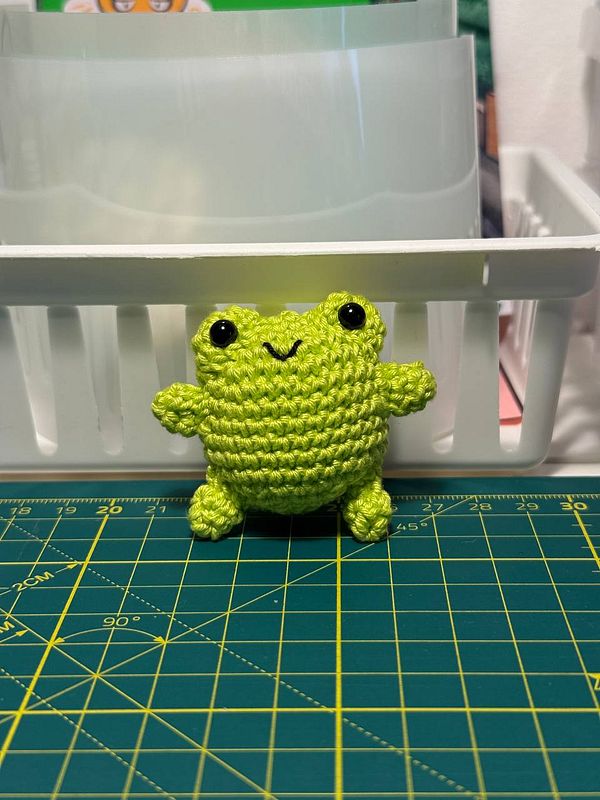 A crocheted green critter with a cheerful expression is placed on a cutting mat.