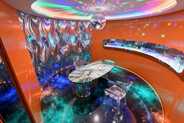 A circular home office with curved orange walls features an oval desk with a glass top and marble-like surface, flanked by two transparent acrylic chairs on a glossy galaxy-patterned floor. A mirrored disco ball and cove ceiling lights cast multicolored reflections onto a wavy iridescent chrome wall and a long mirrored display niche, creating a vibrant, space-age atmosphere.