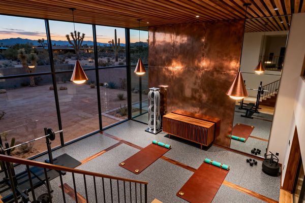 A mid-century modern home gym with floor-to-ceiling black-framed windows overlooks a desert landscape at dusk. Inside, a patinated copper wall and matching cone pendants glow beside a slatted walnut ceiling and credenza. Speckled gray rubber flooring with wood inlay frames two cognac leather exercise mats with teal bolsters. A full-height mirror reflects the space, while a squat bench, dumbbells, weight plates, and a chrome rope stand complete the workout area.