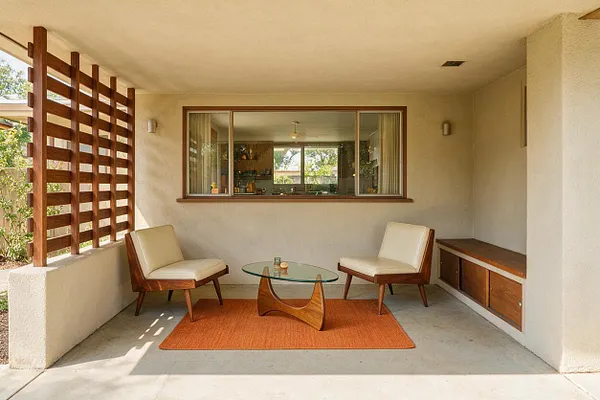 A covered front porch in mid-century modern style features two low lounge chairs with cream cushions and walnut wood frames, facing a sculptural glass coffee table on an orange rug. A built-in wood bench lines the right wall, while a horizontal slatted wood screen on the left filters sunlight and adds privacy. A large window and neutral stucco walls complete the calm, open-air space.