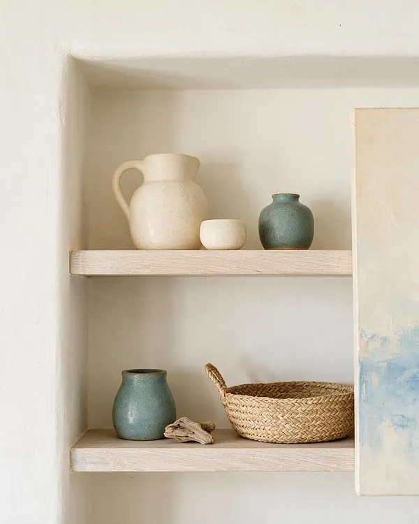 A recessed wall niche with two light wood shelves displays cream ceramic vessels, small blue-green pottery, a woven basket, and a small piece of driftwood. The palette is soft and airy, with white plaster walls and a serene coastal feel.