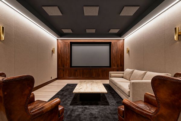 Transitional home theater with a large screen set into floor-to-ceiling walnut cabinets on the far wall. Neutral fabric-clad acoustic wall panels line both sides, with a beige sofa, two cognac leather armchairs, and a travertine coffee table on a charcoal rug. Perimeter LED cove lighting and brass sconces illuminate pale wood flooring and a dark ceiling dotted with acoustic pads, creating a warm, balanced space for immersive viewing.