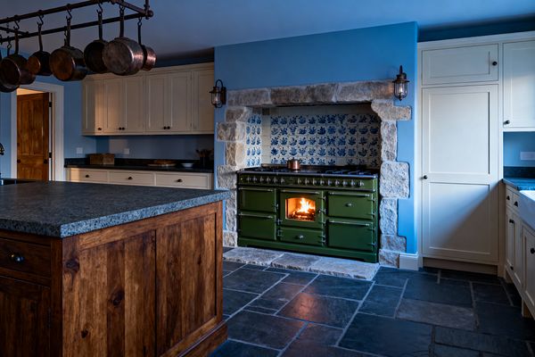 A farmhouse kitchen with dusty blue walls and cream Shaker cabinets centers on a deep green enamel range burning behind a glass door, set within a chunky stone-framed alcove. The niche is lined with blue-and-white patterned tiles, while dark granite counters, a stained wood island, an iron pot rack with hanging copper pans, a white apron-front sink, slate flagstone flooring, and two metal-caged sconces complete the warm, rustic scene.
