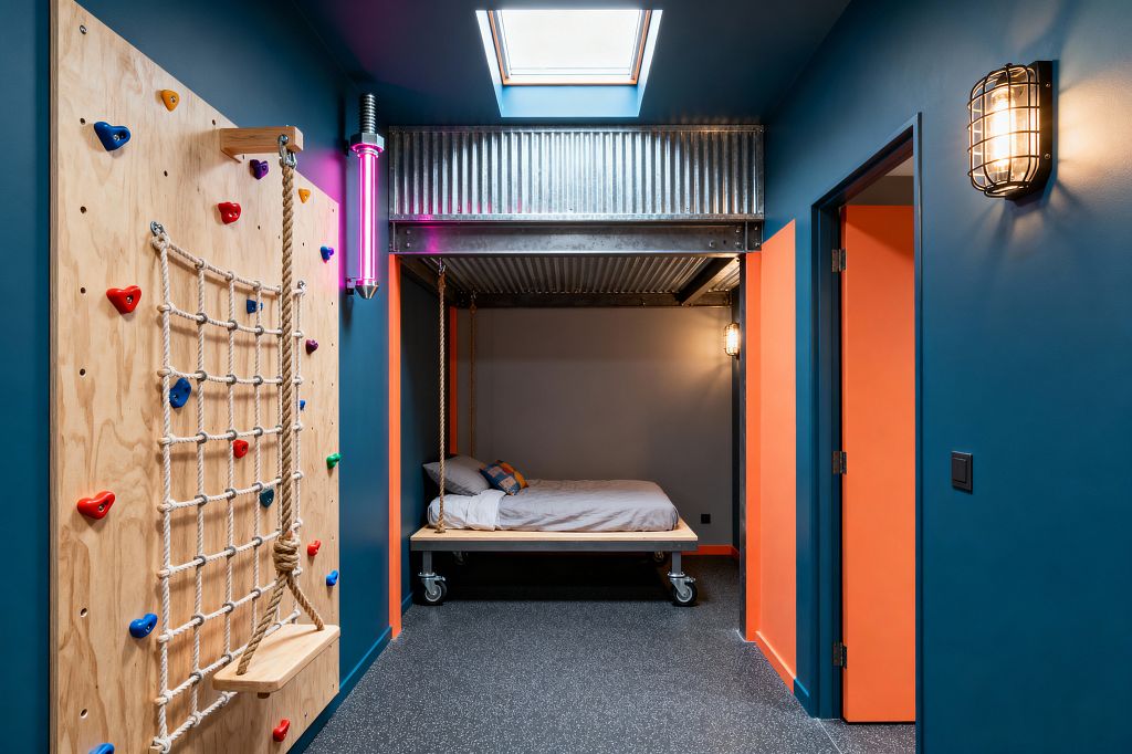A kids’ room in industrial style features deep teal walls with bright orange doors, a central skylight, and caged wall sconces. A gray alcove holds a rope-suspended platform bed on casters beneath a corrugated metal canopy, while a plywood climbing wall with multicolored grips, a rope net, and a small rope swing lines the opposite wall; a neon pink tube light and speckled rubber flooring complete the playful, durable space.