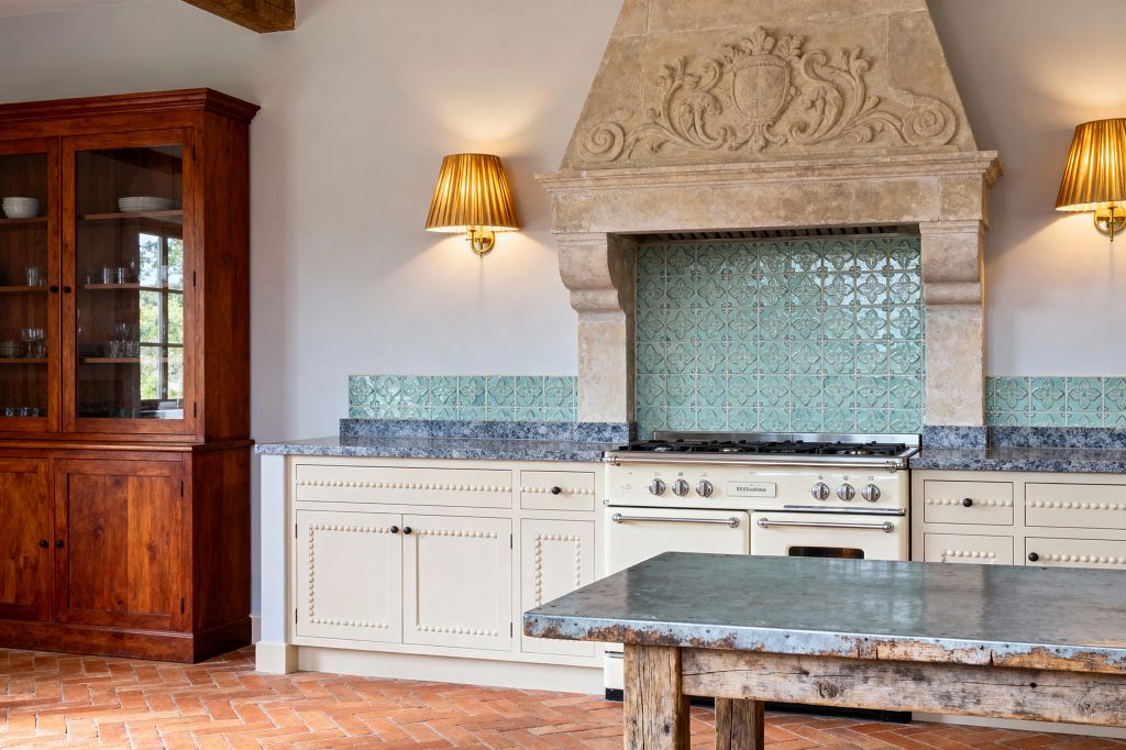 A traditional kitchen features a cream enamel range set beneath a grand carved limestone hood. Glossy seafoam tiles with a raised motif line the backsplash above blue-gray granite counters and cream cabinets detailed with bead molding and dark knobs. Brass wall sconces with pleated gold shades flank the hood. At left stands a tall glass-front hutch in warm wood. The floor is herringbone terracotta, and a rustic table with a weathered zinc top sits in front.