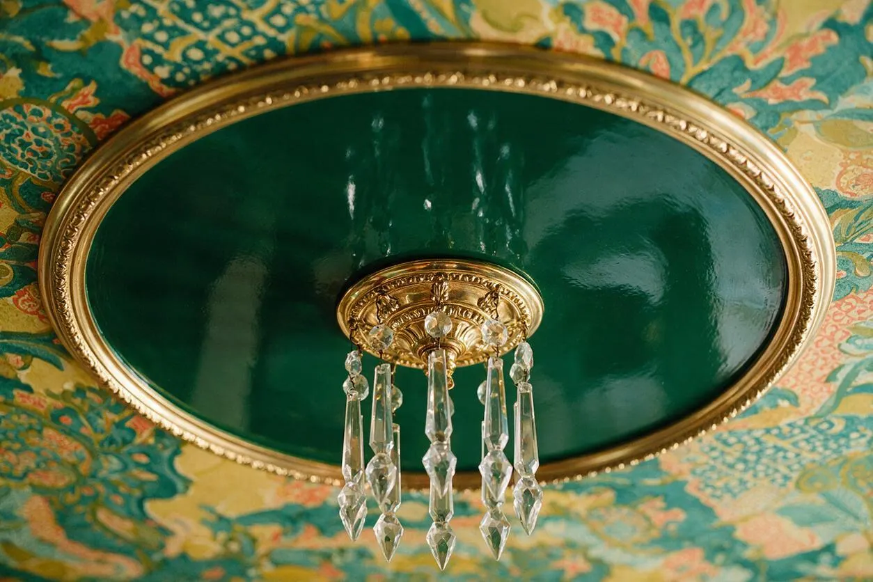 A close-up view of an ornate ceiling detail featuring a glossy emerald green oval medallion framed in gold, with a small crystal chandelier hanging from the center. Around it, the ceiling is covered in a vibrant multicolor patterned wallpaper with teal, gold, coral, and cream motifs.