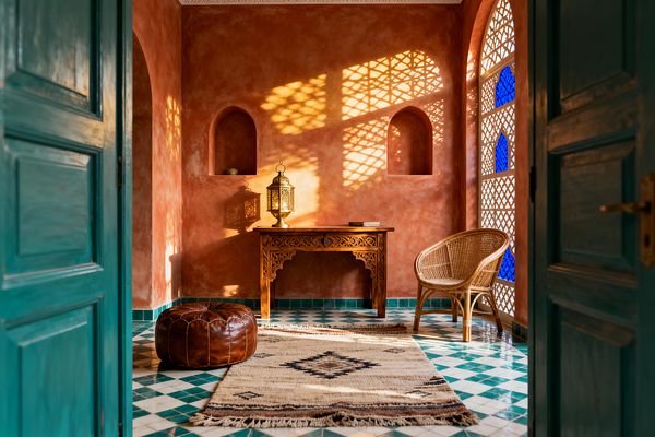 View through teal doors into a sunlit Moroccan-style home office with terracotta tadelakt walls and two arched niches. A carved wooden writing desk holds a brass lantern, while a rattan chair sits by an arched lattice window inset with cobalt glass, casting patterned shadows; a brown leather pouf and a fringed Berber rug rest on green-and-white zellige tiles, with minimal decor and warm afternoon light creating a calm atmosphere.