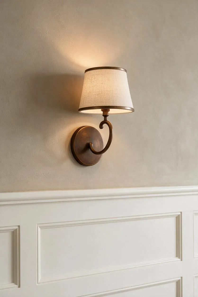 A transitional wall sconce with a curved bronze arm and round backplate is mounted on a warm beige wall above white wainscoting. The small tapered linen shade casts a soft glow, creating a classic and elegant accent.