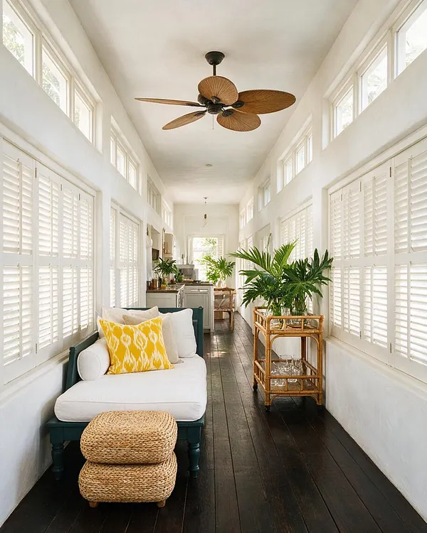A long, bright sunroom with white walls and plantation shutters on both sides, dark wood floors, and high clerestory windows. The space features a teal-framed white sofa with yellow and cream pillows, woven ottomans, a bamboo bar cart, lush green potted palms, and a tropical ceiling fan with palm-leaf blades.