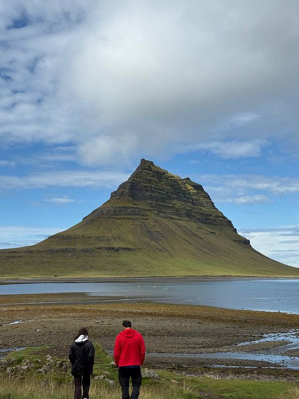 Two people stand by a body of water, gazing at the iconic Kirkjufell mountain under a partly cloudy sky.