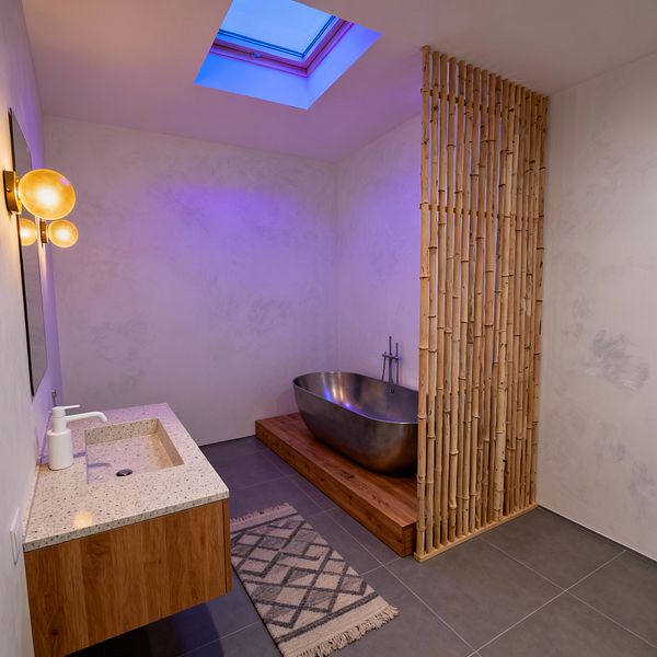 A minimalist Scandinavian bathroom features pale plaster walls and large gray floor tiles under a skylight that casts cool light. At the back, a brushed-metal freestanding tub sits on a raised wood platform next to a vertical bamboo slat partition. On the left, a floating wood vanity with a speckled terrazzo countertop and integrated sink is paired with a simple white faucet, a rectangular mirror, and a warm double-globe wall sconce. A small patterned runner lies on the floor.