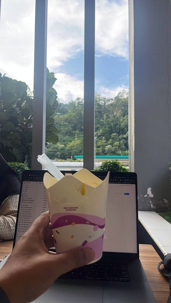A hand holds a cup of durian ice cream in front of a laptop and a scenic view.