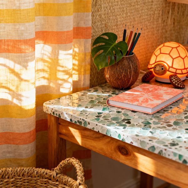 A close-up of a tropical-themed kids’ study nook shows a terrazzo tabletop with green sea-glass chips on a honey wood base. A coconut shell pencil cup with a small monstera leaf and colored pencils sits beside a glowing turtle nightlight and a coral tie-dye notebook, while sunlight filters through yellow and orange striped linen curtains, casting leafy shadows across woven seagrass wallcovering and a rattan basket under the desk.