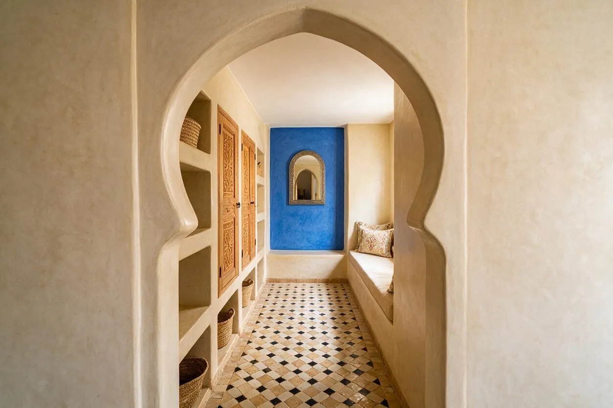 A narrow Moroccan-style closet with a sculpted horseshoe arch framing the entrance, built-in niches and carved wooden cabinet doors on the left, and a cushioned bench with patterned pillows on the right. The space ends with a vivid blue accent wall, an ornate mirror, woven baskets, and a geometric tiled floor in warm neutral tones.
