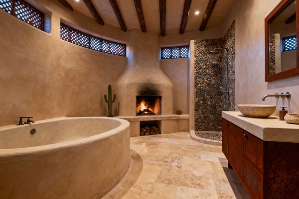 A Southwestern bathroom features curving stucco walls centered on a kiva-style fireplace with stacked firewood and a glowing flame. A rounded built-in soaking tub with dark bronze fixtures sits at left, while a walk-in shower at right is lined with river pebbles and a rainfall head. Travertine tiles cover the floor, dark timber beams span the ceiling, and a wood vanity with stone vessel sinks and a rust-toned framed mirror completes the space, with lattice clerestory windows and a cactus accent.