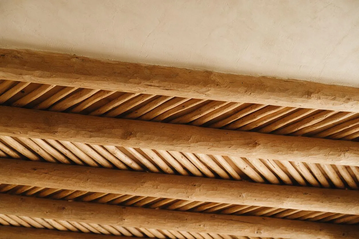 An angled view looking up at a ceiling with thick rustic wood beams and narrow diagonal wood slats between them. The warm honey-brown wood contrasts with a smooth pale plaster ceiling, creating a Southwestern-inspired architectural detail.