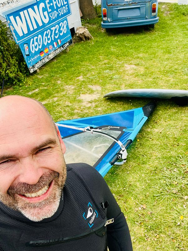 A man in a wetsuit smiles at the camera while standing next to windsurfing equipment and a vintage blue van.