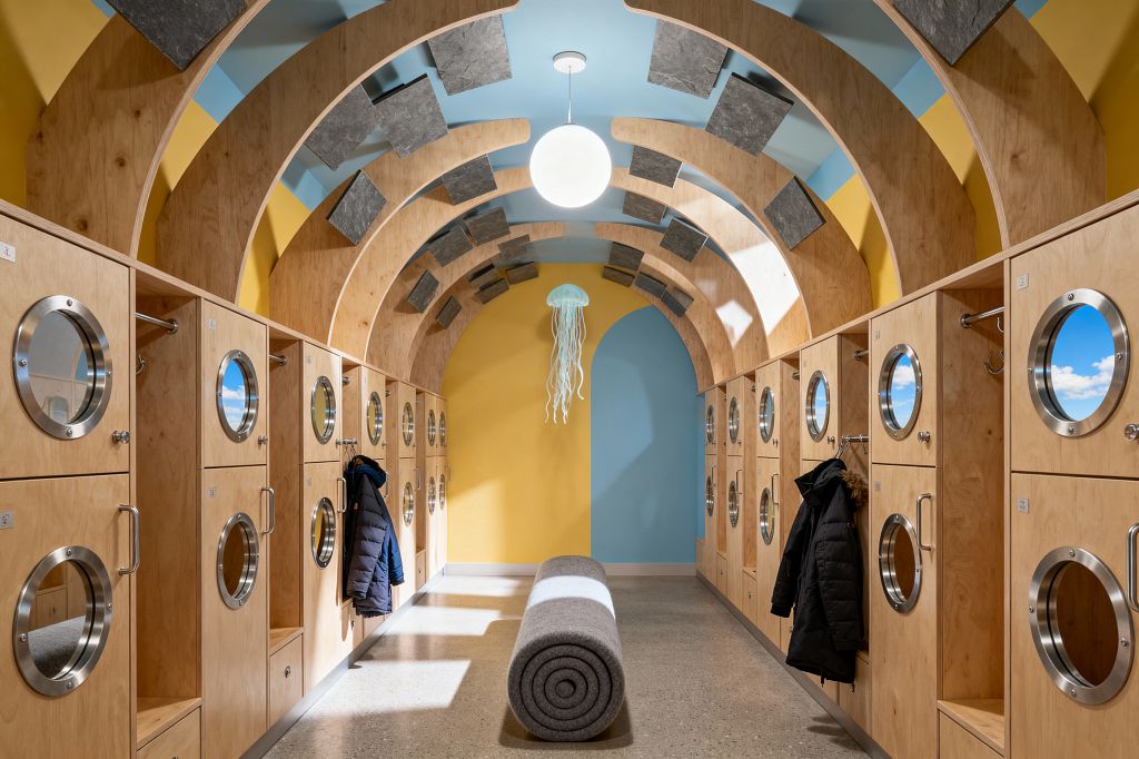 Long, tunnel-like mudroom with curved plywood arches overhead and walls split between sunny yellow and sky blue. Light wood lockers line both sides with round stainless porthole doors showing cloud imagery, plus hooks and handles; a rolled gray felt bench rests on polished concrete below a glowing globe and a jellyfish-like pendant, while rectangular gray acoustic panels appear to float across the ceiling, creating a playful, surreal atmosphere.