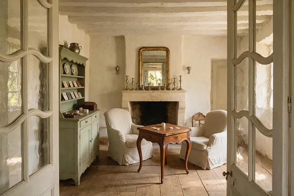 A cozy French Country game room with two upholstered armchairs facing a small carved wooden game table in front of a stone fireplace. A painted green cabinet with cards and dice sits to the left, while distressed wood floors, exposed ceiling beams, and open glass-paneled doors create a soft, rustic atmosphere.