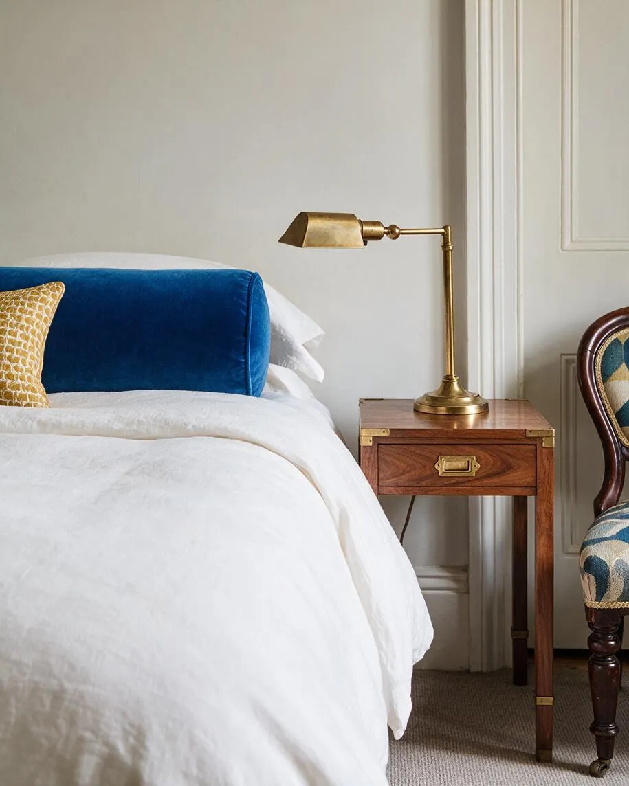 A refined bedroom vignette shows a bed dressed in white bedding with a deep blue velvet bolster pillow and a patterned gold accent pillow. Beside it, a polished brass task lamp sits on a small walnut-toned wooden nightstand with brass corners and a drawer pull, next to a partial view of a patterned upholstered chair and paneled wall.