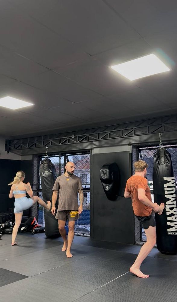 A man practicing Muay Thai in a gym ring throwing a high kick at an opponent in red shorts.