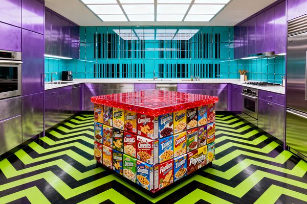 A U-shaped kitchen in bold Pop Art colors features glossy purple cabinets, white countertops, and a turquoise tile backsplash beneath a luminous grid ceiling. At the center stands a cube island wrapped in vintage-style cereal box graphics with a red resin top, set on a lime-green and black zigzag floor; stainless steel oven, cooktop, dishwasher, and refrigerator line the perimeter with under-cabinet lighting illuminating the work surfaces.