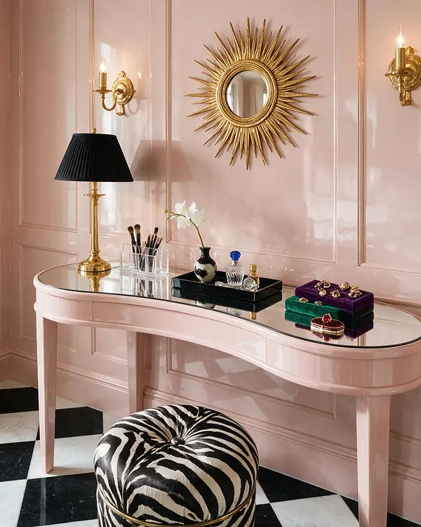 A glamorous vanity vignette with a glossy blush pink console table topped with a mirrored surface, a black-shaded brass lamp, makeup brushes, small perfume bottles, and colorful jewelry boxes. The wall has pale pink paneling with gold trim, a sunburst mirror, and brass sconces, while a black-and-white checkered floor and zebra-print ottoman add bold contrast.