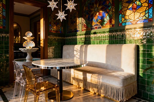 A jewel-toned patio features stained-glass windows over glossy emerald green tiles. A cream velvet banquette with bullion fringe lines one wall beside a round marble table with a brass-edged pedestal base. Clear and amber acrylic chairs sit around the table. Overhead, three star-shaped crystal pendants hang from brass chains. A black-and-white marble floor grounds the space, while a tiered shell fountain is visible through a wooden doorway, all bathed in warm late-afternoon light.