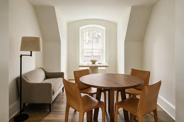 A narrow transitional game room with a round wooden table surrounded by four tan leather chairs, a gray upholstered settee, and a black floor lamp with a beige shade. The room features white walls, warm wood flooring, and a bright arched window at the far end with a small stone-topped console beneath it.