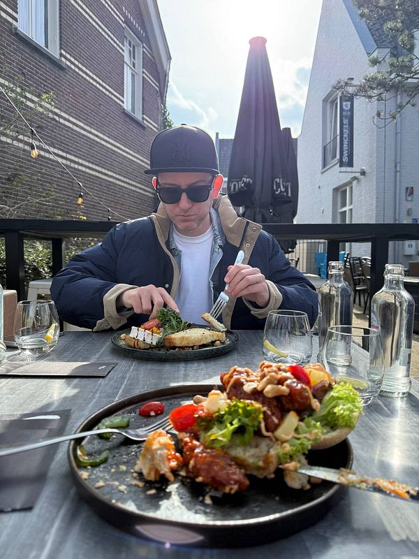 Portrait of a man with a beard and slicked-back hair, wearing a brown jacket and a white collared shirt, sitting indoors with a thoughtful expression.