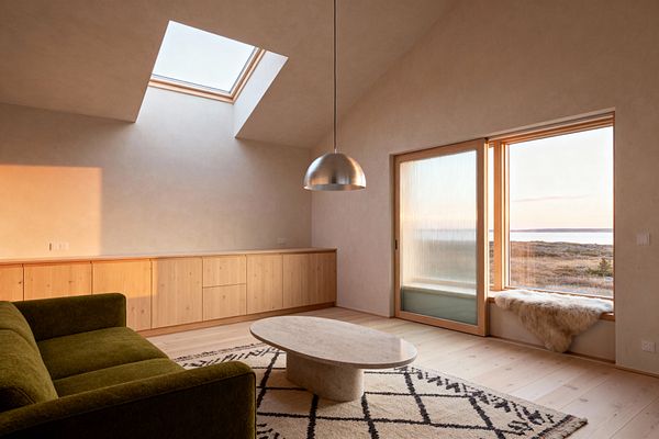 A Scandinavian-style living room with a vaulted ceiling and skylight features a low olive green sofa facing an oval light-stone coffee table on a cream wool rug with a black diamond pattern. A long run of knotty pine cabinetry lines one wall beneath warm, plastered surfaces. A brushed aluminum dome pendant hangs centrally. To the right, a large picture window and a sliding door with fluted glass frame a landscape view, with a sheepskin draped on the window seat and pale wood floors throughout.