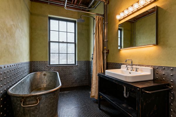 An industrial bathroom with an oval galvanized metal soaking tub set against riveted steel wainscoting and black penny-round floor tiles. Olive-toned plaster walls, an exposed rainfall shower with a canvas curtain, a wire-glass window, and a blackened steel vanity topped with a white ceramic sink sit below a wide mirror lit by warm bulbs and an LED strip, with visible copper and steel pipes throughout.