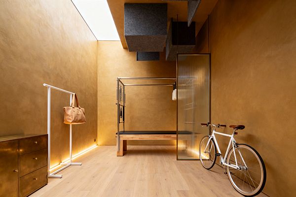 A contemporary home gym with warm ochre plaster walls and pale oak floors centers on a Pilates Cadillac with a stainless frame and wood base. Gray felt acoustic cubes hang from the ceiling near a linear skylight, while concealed LED lighting washes the lower walls. At left is a brass three-drawer cabinet and a slim white clothes rail holding a tan tote; at right a white bicycle rests beside a ribbed glass partition.