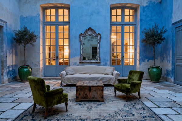 Symmetrical French Country living room set against limewashed blue plaster walls, with a linen slipcovered sofa centered below a distressed carved mirror. Two moss‑green velvet armchairs face a rustic reclaimed‑wood trunk on a faded floral rug, while tall pale‑blue multipane French doors glow with warm light from adjoining rooms; large green glazed urns with small trees flank the scene on aged limestone flagstones.
