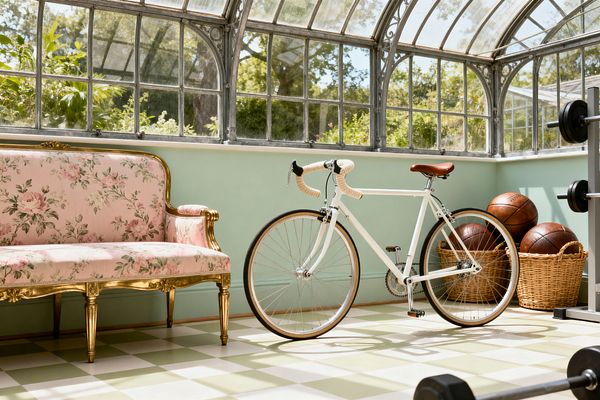 A sunlit conservatory-turned-home gym features a mint wall below arched, patinated metal-and-glass windows. A vintage pink floral settee with a gilded wood frame sits on the left. Centered is a white road bicycle with cream-wrapped handlebars and a tan leather saddle. To the right, wicker baskets hold brown leather basketballs beside a weight rack with black plates and a floor dumbbell. The floor shows sage-and-cream checkerboard tile.