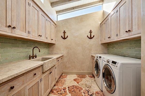 Galley-style laundry room with whitewashed wood Shaker cabinets on both sides, a pale green glass subway tile backsplash, and honed stone countertops with a carved apron-front sink and antique brass bridge faucet. The floor features coral and sand-toned porous stone pavers, sand-colored plaster walls hold two wooden anchor decorations, and a clerestory window with exposed beams brings in natural daylight.