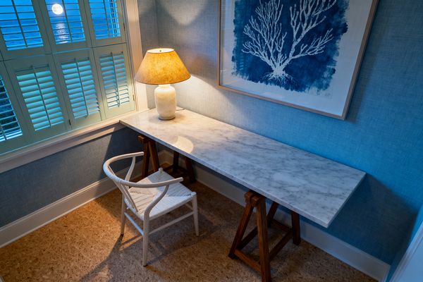 A compact coastal home office features a long white marble slab desk on walnut trestle bases, paired with a white wishbone-style chair with a woven paper-cord seat. Blue grasscloth walls, an indigo coral print in a light wood frame, and a stone table lamp with a warm shade create a calm glow, while pale seafoam plantation shutters frame a window with a moonlit ocean view over sandy-toned cork flooring.