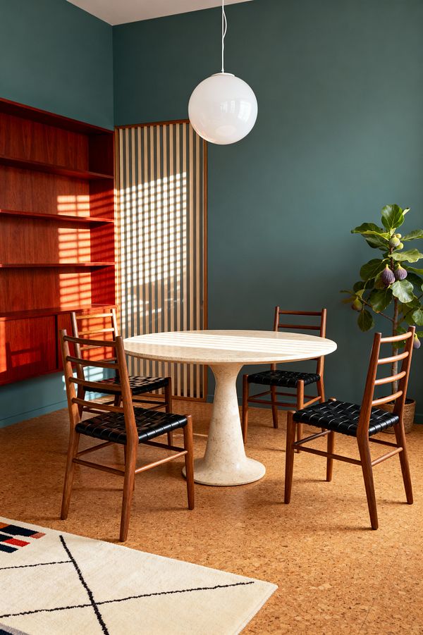 A Mid-Century Modern dining nook features a round travertine pedestal table centered beneath a white globe pendant. Four ladder-back wooden chairs with woven black leather seats surround the table. Matte teal walls contrast with a built-in reddish wood shelving unit, while a vertical slatted screen casts a grid of sunlight across the surfaces. Cork flooring grounds the space, partly layered with a cream rug with fine black lines, and a potted fig tree sits to the right.