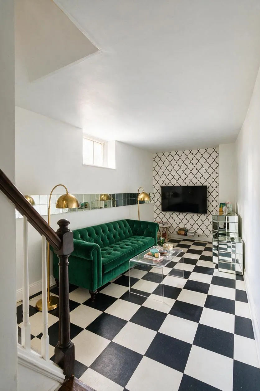 A stylish basement lounge with a black-and-white checkered floor, a deep green tufted velvet sofa, and brass arc floor lamps. The far wall features patterned wallpaper with a wall-mounted TV, mirrored storage, and a clear acrylic coffee table, all lit by a small high window.