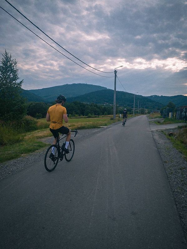 Two cyclists riding on a rural road surrounded by hills and cloudy skies.